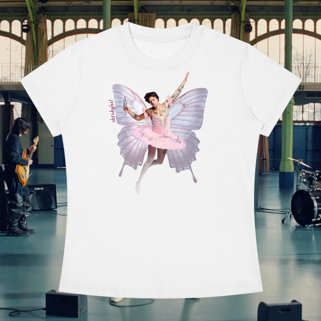 A white short-sleeve crewneck t-shirt featuring a graphic print of a ballerina with butterfly wings in a pink tutu, displayed against a blurred indoor stage background with a band and instruments.