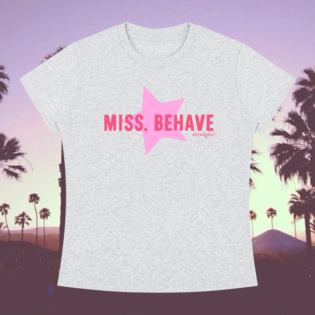 A light heather gray short-sleeve t-shirt with a pink graphic on the chest featuring a large pink star and the text 'MISS. BEHAVE' in pink, with a small 'missbehavestreet' brand mark underneath.